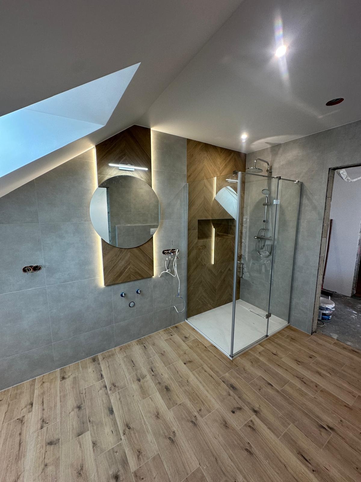 Wykończona łazienka na poddaszu: kabina prysznicowa, okrągłe lustro z podświetleniem LED, drewnopodobne płytki na podłodze i ścianie z wzorem jodełki. Częściowo widoczne kable.