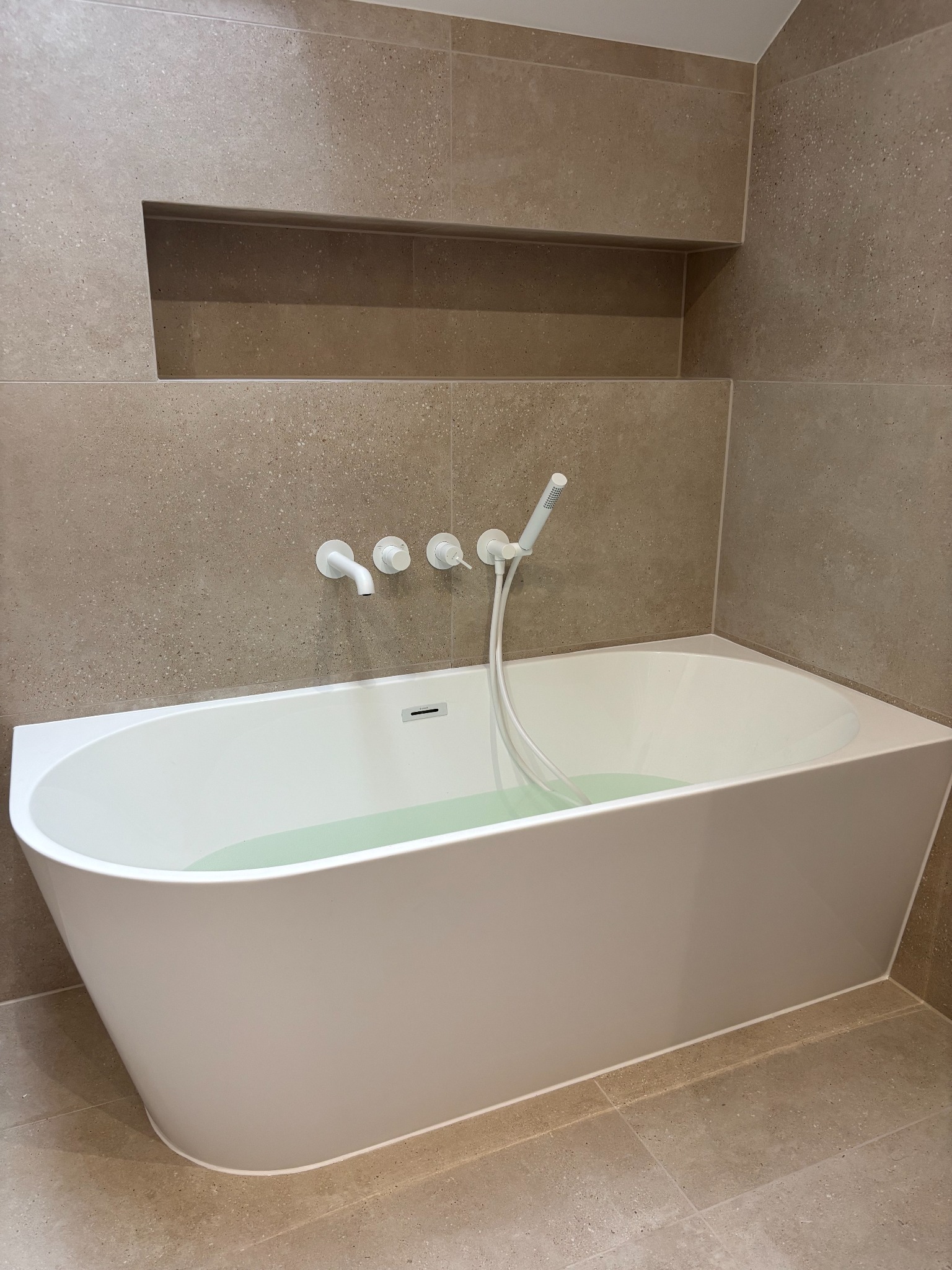 Nowoczesna łazienka z beżowymi płytkami i białą wanną wypełnioną wodą. W ścianie wnęka, armatura w kolorze białym. Minimalistyczny design.