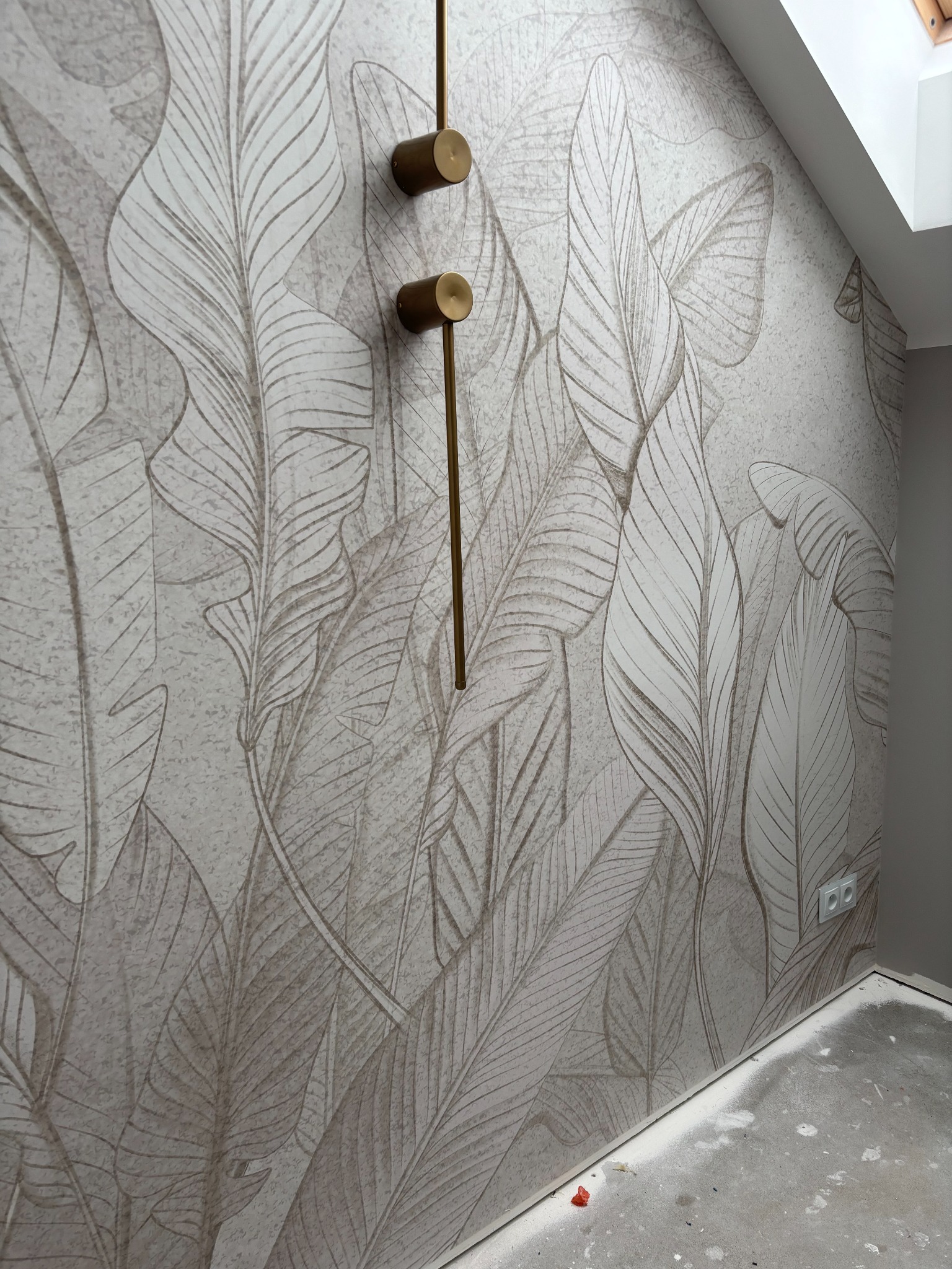 Ściana z tapetą w liście i złotymi lampami ściennymi. Widoczny fragment sufitu i gniazdko elektryczne. Podłoga w trakcie wykończenia.