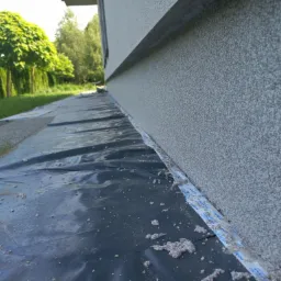 Szara elewacja budynku z marmolitu, zabezpieczona folią ochronną i taśmą przy gruncie.