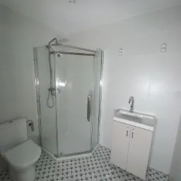 Wykończona łazienka z kabiną prysznicową narożną, toaletą i szafką z umywalką na wzorzystej podłodze, oświetlenie sufitowe