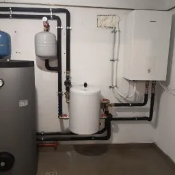 Kotłownia z szarym zasobnikiem ciepłej wody, białym zbiornikiem wyrównawczym i wiszącym białym piecem Panasonic, rury w izolacji, widoczne zawory i manometr.