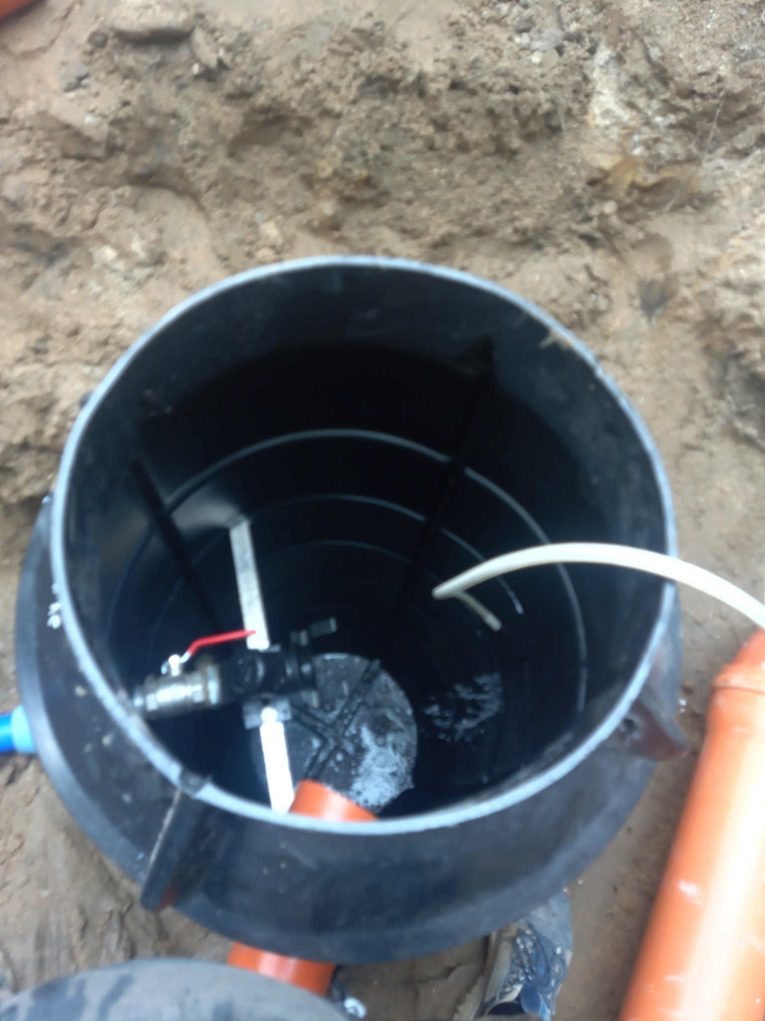 Czarny zbiornik przydomowej oczyszczalni ścieków w wykopie, widoczne rury, zawór i woda.