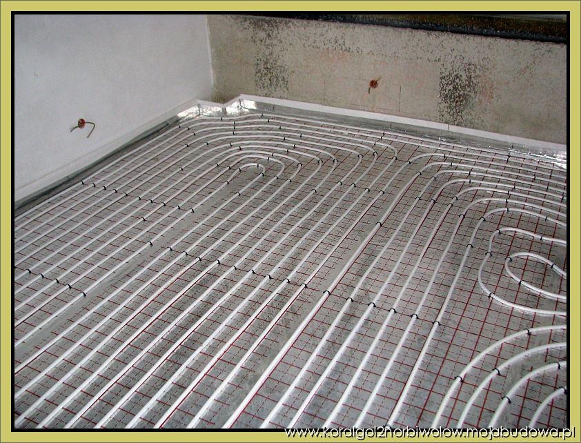 Białe rury ogrzewania podłogowego ułożone w pętle na szarej siatce z czerwonymi liniami, widok z góry na surowy beton i fragment ściany.