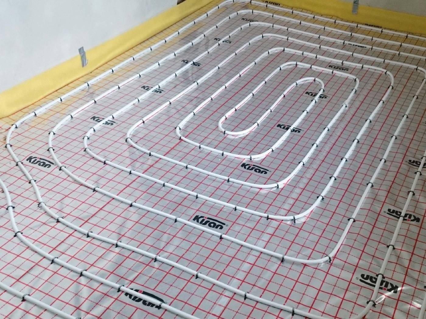 Rozłożona instalacja ogrzewania podłogowego z rurkami marki Kisan na folii z czerwoną siatką, widoczne mocowania rur do podłoża i żółta izolacja przy ścianie.