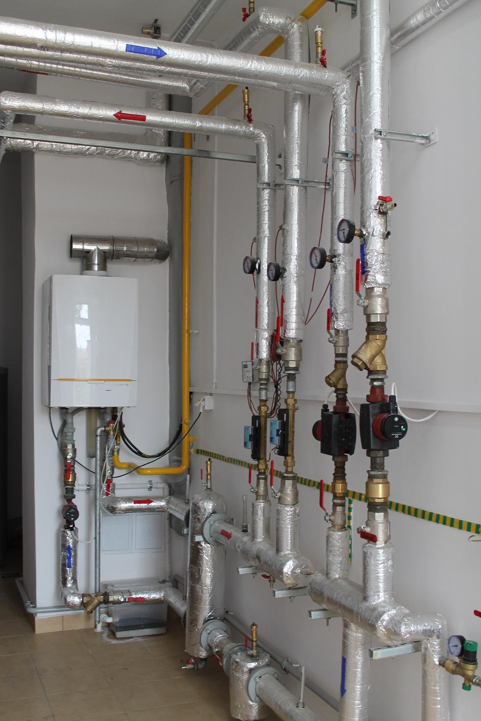 Biały piec gazowy kondensacyjny z aluminiowym kominem, żółte rury gazowe i rzędy izolowanych srebrną folią rur z zaworami, manometrami i pompami obiegowymi w kotłowni z płytkami na podłodze.