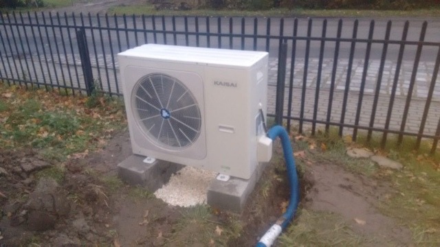 Zewnętrzna jednostka pompy ciepła marki Kaisai, zamontowana na betonowych bloczkach przy metalowym ogrodzeniu, z widocznym niebieskim wężem odprowadzającym skropliny.