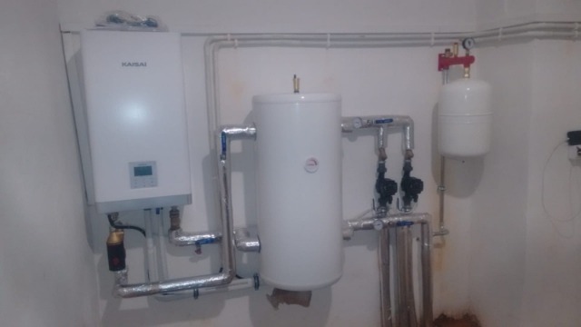 Biały piec gazowy Kaisai, zbiornik wody i naczynie przeponowe połączone rurami w pomieszczeniu kotłowni, widoczne zawory i manometry.