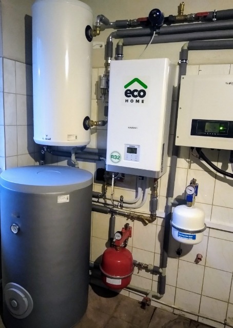 Zestaw urządzeń grzewczych w kotłowni: zbiornik akumulacyjny szary, bojler biały, pompa ciepła z logo Eco Home, naczynia wzbiorcze czerwone i białe, rury i zawory.