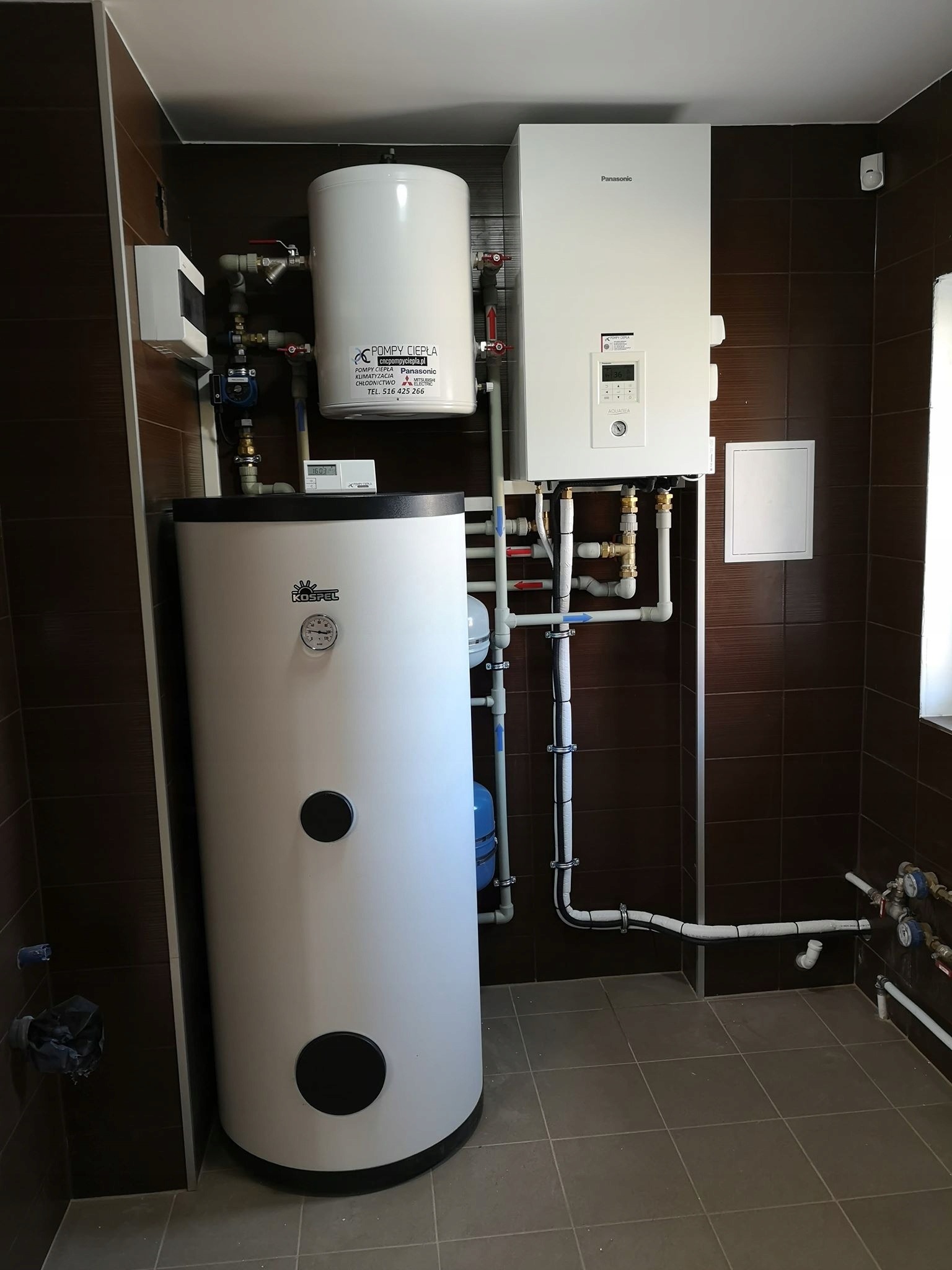 Kompletna instalacja pompy ciepła z zasobnikiem ciepłej wody użytkowej i jednostką wewnętrzną Panasonic Aquarea, widoczne rury i zawory, brązowe płytki na ścianach i szare na podłodze.