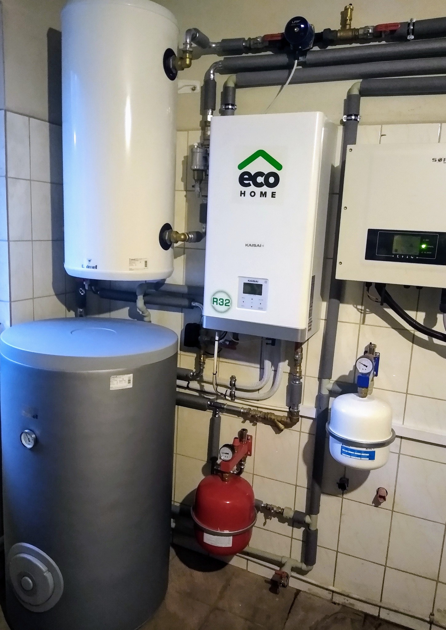 Zestaw urządzeń grzewczych w kotłowni: biały bojler, szary zbiornik akumulacyjny, biała pompa ciepła Kaisai z logo Eco Home, oraz czerwone naczynie przeponowe, połączone rurami w pomieszczeniu...