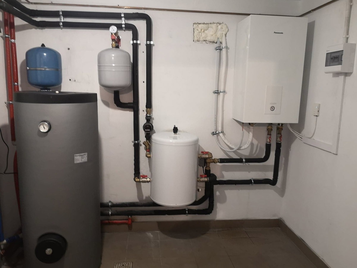 Kotłownia z szarym zasobnikiem ciepłej wody, białym zbiornikiem wyrównawczym i wiszącym białym piecem Panasonic, rury w izolacji, widoczne zawory i manometr.