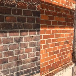 K&M Stolarstwo i Rzemiosło z Pasją Kachel Marcin - Częściowo oczyszczona ceglana ściana budynku, z wyraźnym kontrastem między ciemniejszą, zabrudzoną powierzchnią po lewej i jaśniejszą, odnowioną po prawej, oświetloną przez słońce rzucające cienie...