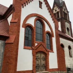 Odnowiona fasada kościoła z czerwonej cegły i białymi elementami, widoczne okna łukowe i drewniane drzwi z metalowymi zdobieniami, częściowo otynkowany fragment ściany z boku.