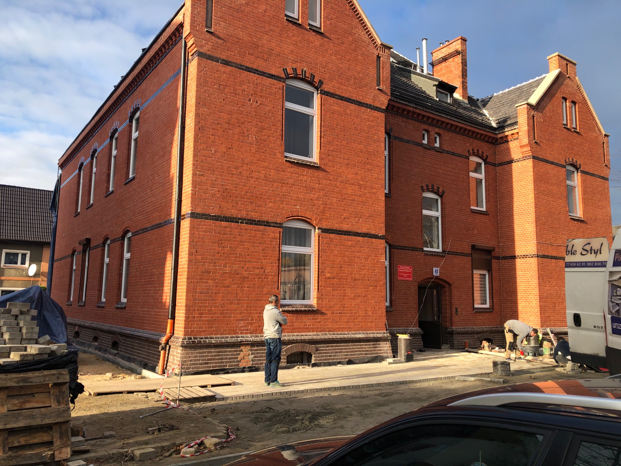 Odrestaurowana, ceglana fasada budynku wielorodzinnego z nową elewacją, widoczny fragment chodnika z kostki, pracownicy w trakcie układania, zaparkowany samochód i częściowo widoczny samochód...
