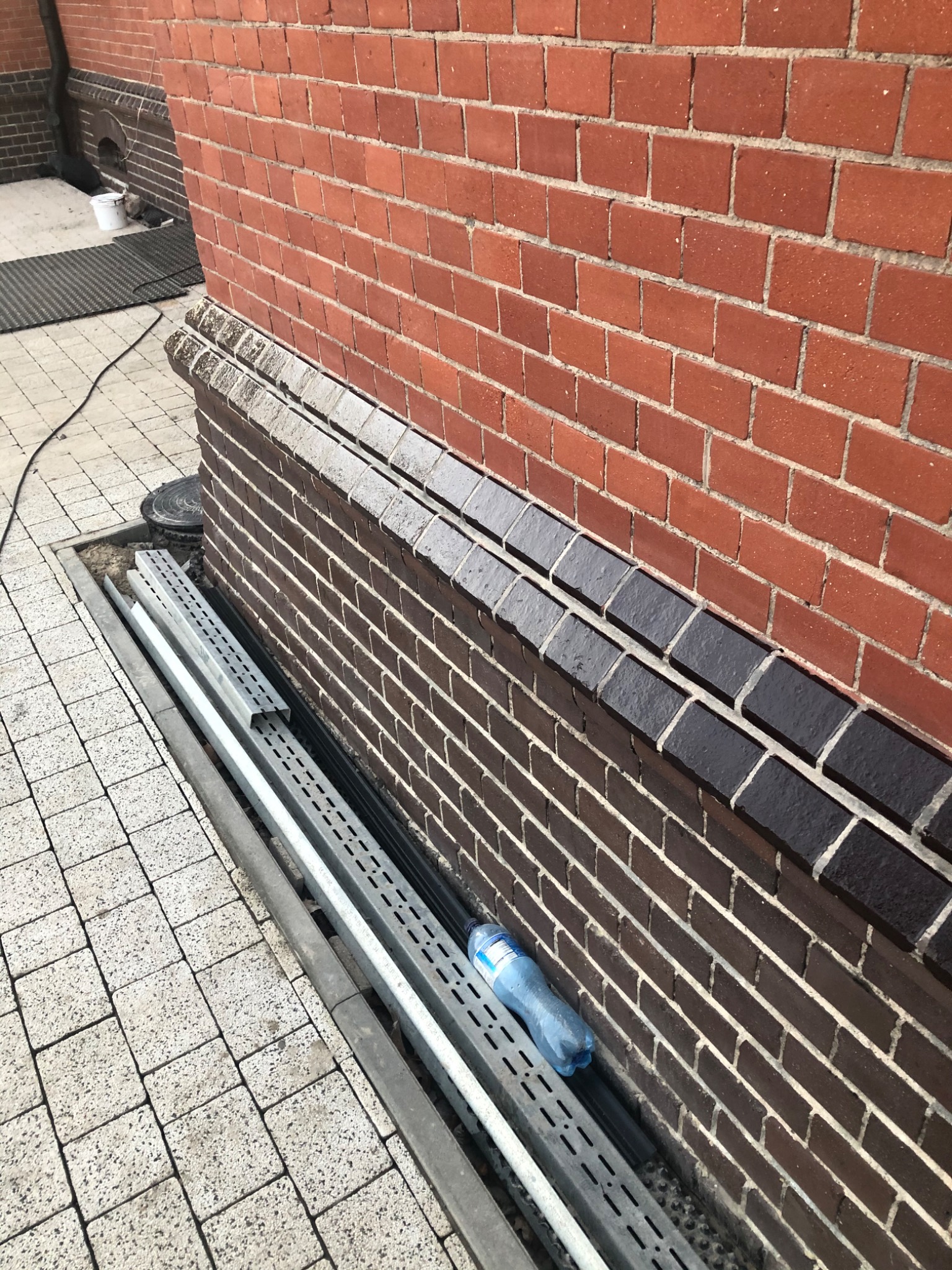 Fragment muru z czerwonej cegły z ciemniejszym pasem pomalowanych cegieł, obok leżą metalowe profile i butelka wody
