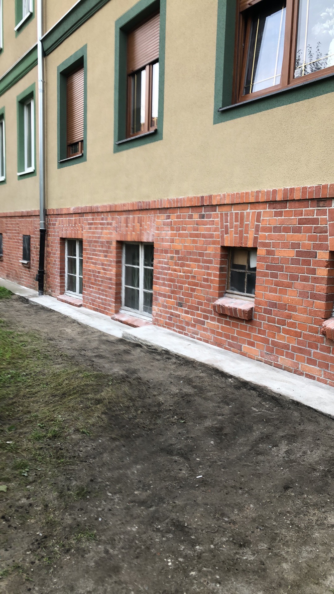 Odnowiona elewacja budynku z ceglanym cokołem, świeżo pomalowana na beżowo górna część z zielonymi obramowaniami okien, widoczne ślady prac ziemnych przy fundamencie.