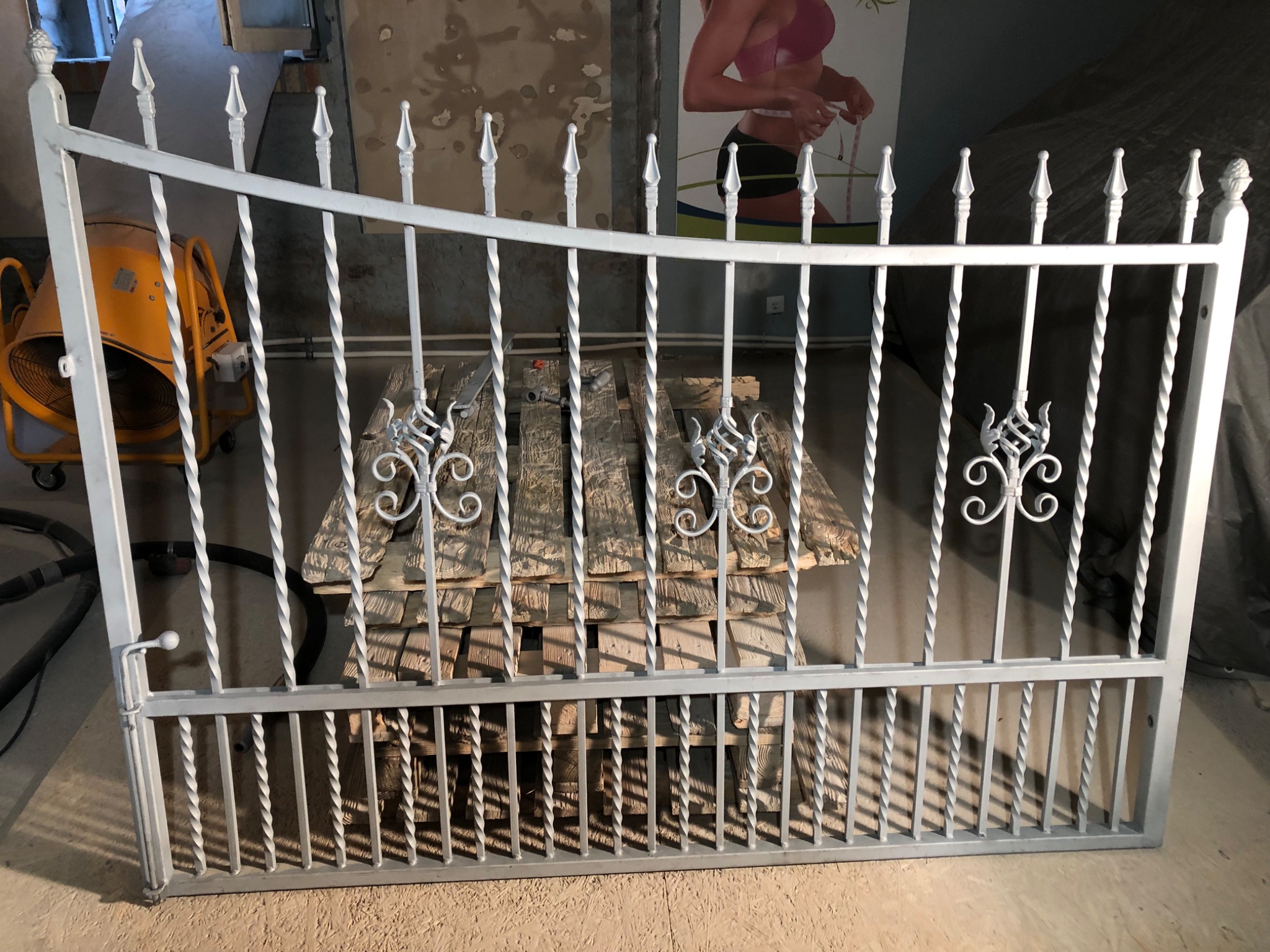 Biała, metalowa furtka z ozdobnymi elementami, oparta o stos pomalowanych desek; w tle widoczna betoniarka i plakat z sylwetką kobiety.