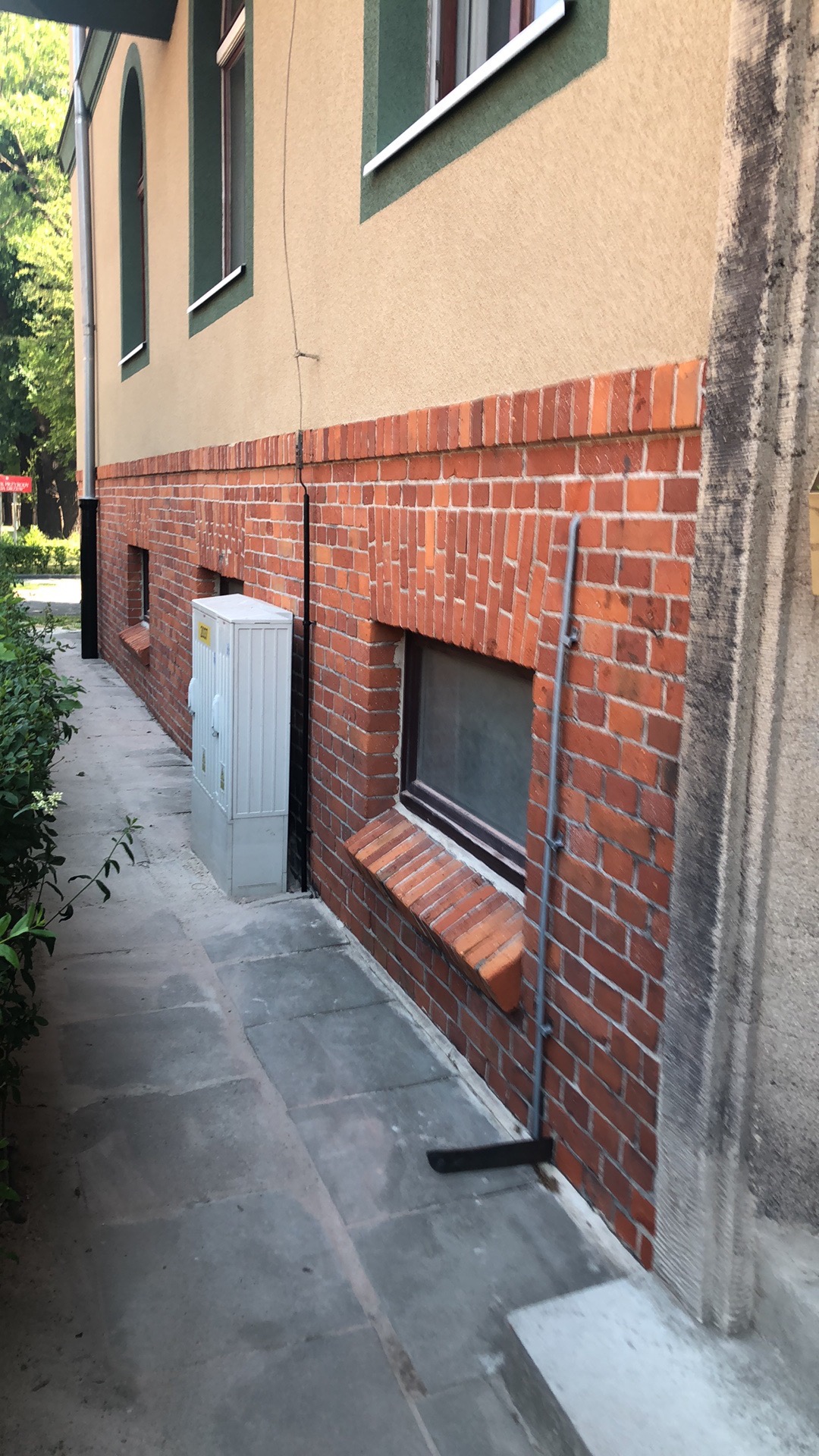 Ceglana podmurówka budynku z oknem piwnicznym i szarą elewacją powyżej, widoczna instalacja przy ścianie i skrzynka elektryczna.