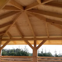 Wnętrze drewnianej altany z widoczną konstrukcją dachu i ścianami z desek, na tle zieleni drzew iglastych.