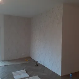 Pokój po zakończonym tapetowaniu, widoczne tapety o delikatnej strukturze, parkiet ułożony w jodełkę, zabezpieczona podłoga, narzędzia i butelki wody na podłodze.