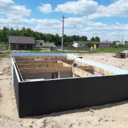 Fundament domu w trakcie budowy, z widocznym szalunkiem i izolacją, na tle piaszczystego terenu i zabudowań jednorodzinnych pod błękitnym niebem.