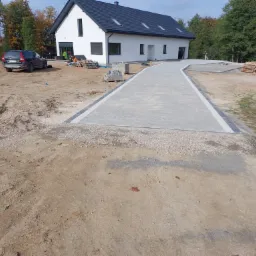 Nowoczesny dom jednorodzinny z jasną elewacją i ciemnym dachem, z nowo wyłożonym chodnikiem z szarej kostki prowadzącym do wejścia, na działce w trakcie zagospodarowywania, z widocznymi paletami...