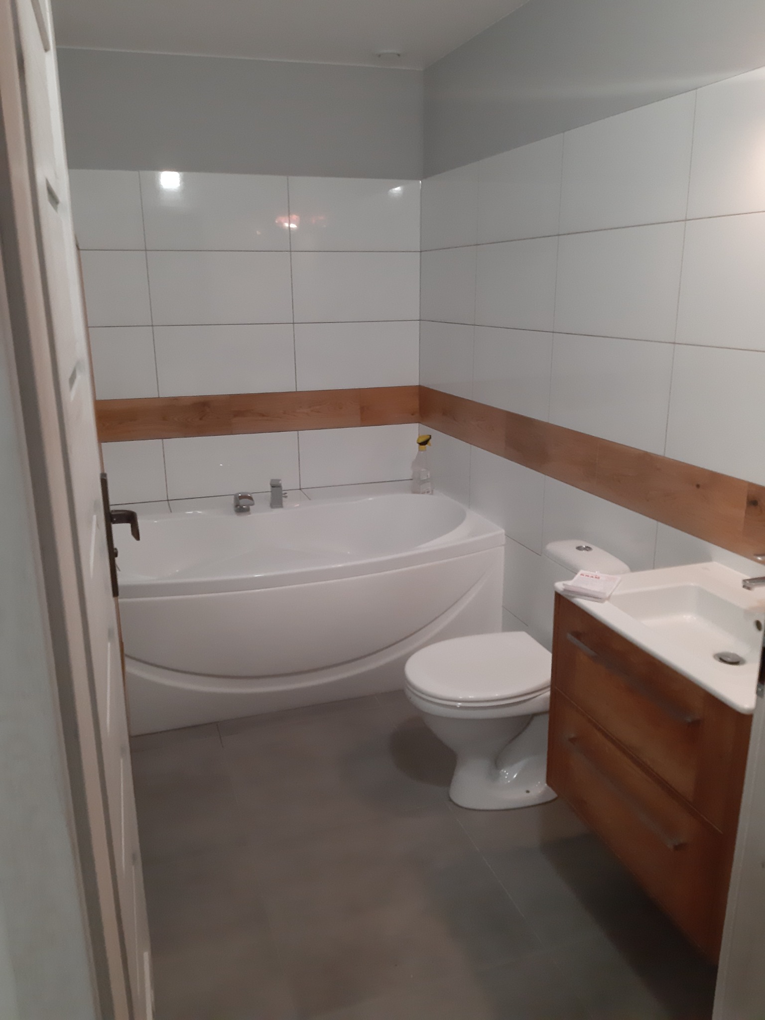 Wykończona łazienka z białą wanną narożną, toaletą i umywalką z drewnianą szafką, białe płytki z drewnianym akcentem, szara podłoga.