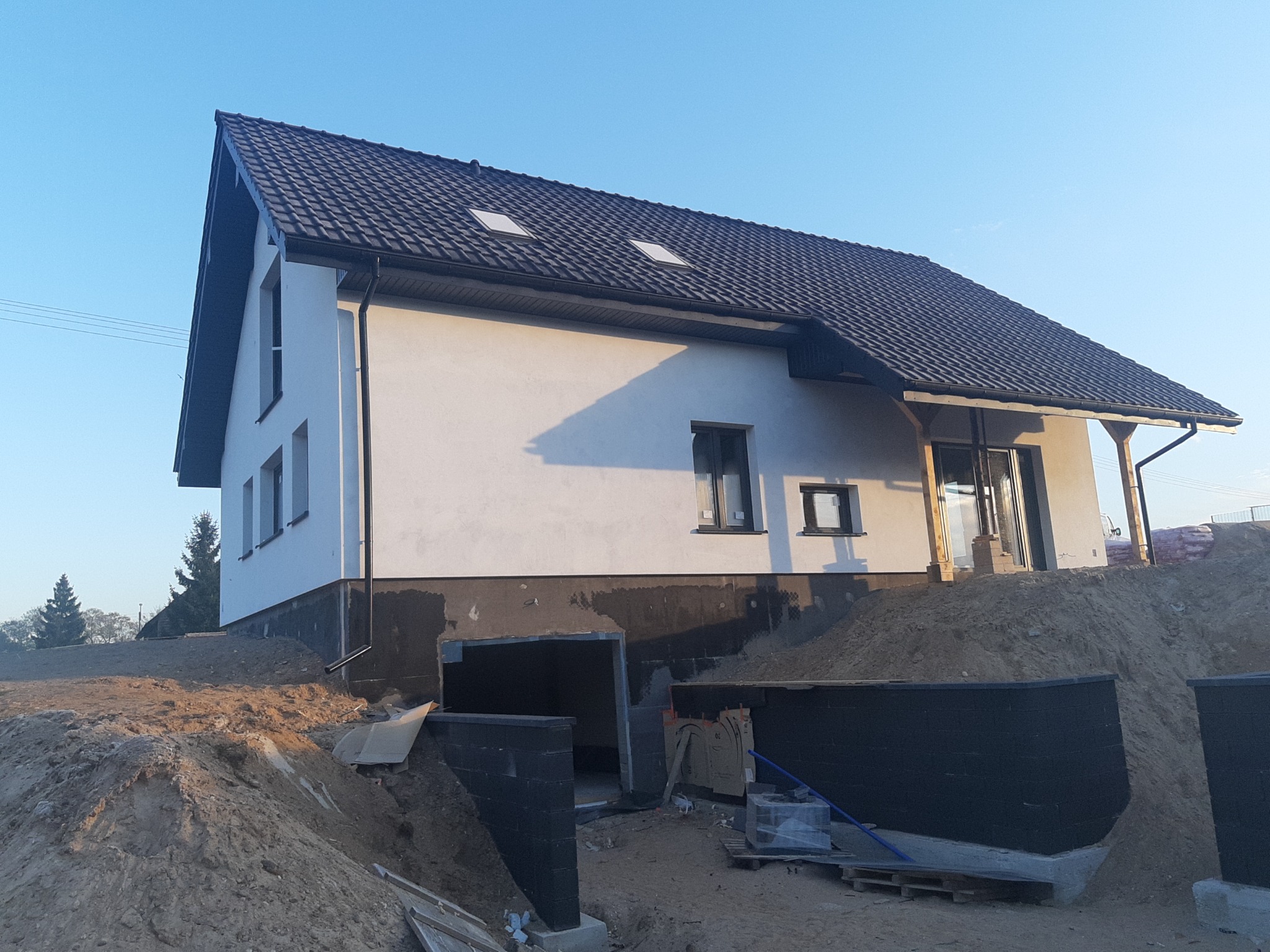 Nowo wybudowany dom jednorodzinny z białymi ścianami i ciemną dachówką, widoczne przygotowania do zagospodarowania terenu wokół budynku, w tym pryzmy ziemi i fundamenty podjazdu.