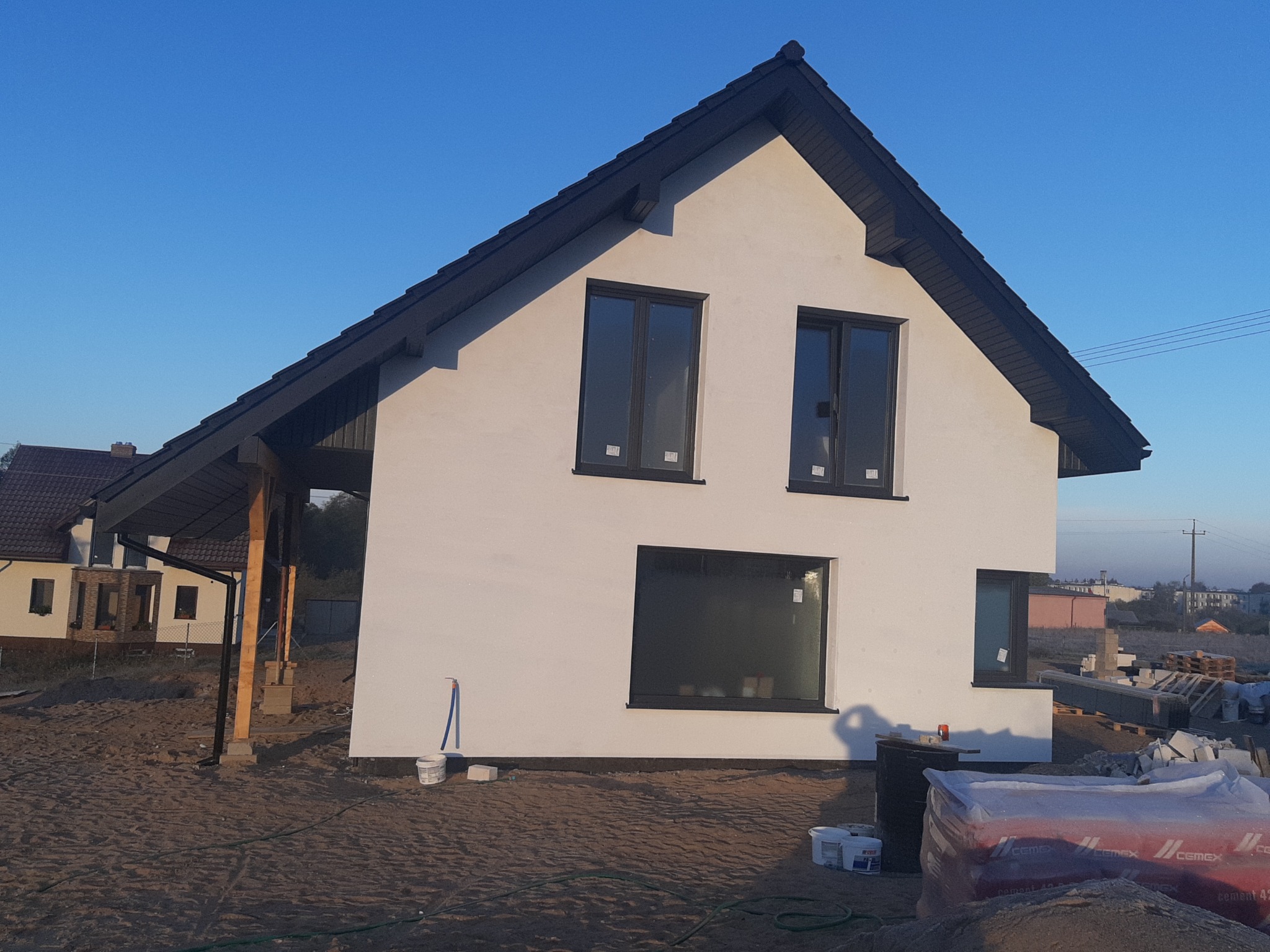 Świeżo otynkowany dom jednorodzinny z ciemną stolarką okienną i dachem, widoczne worki z cementem i narzędzia malarskie na posesji.