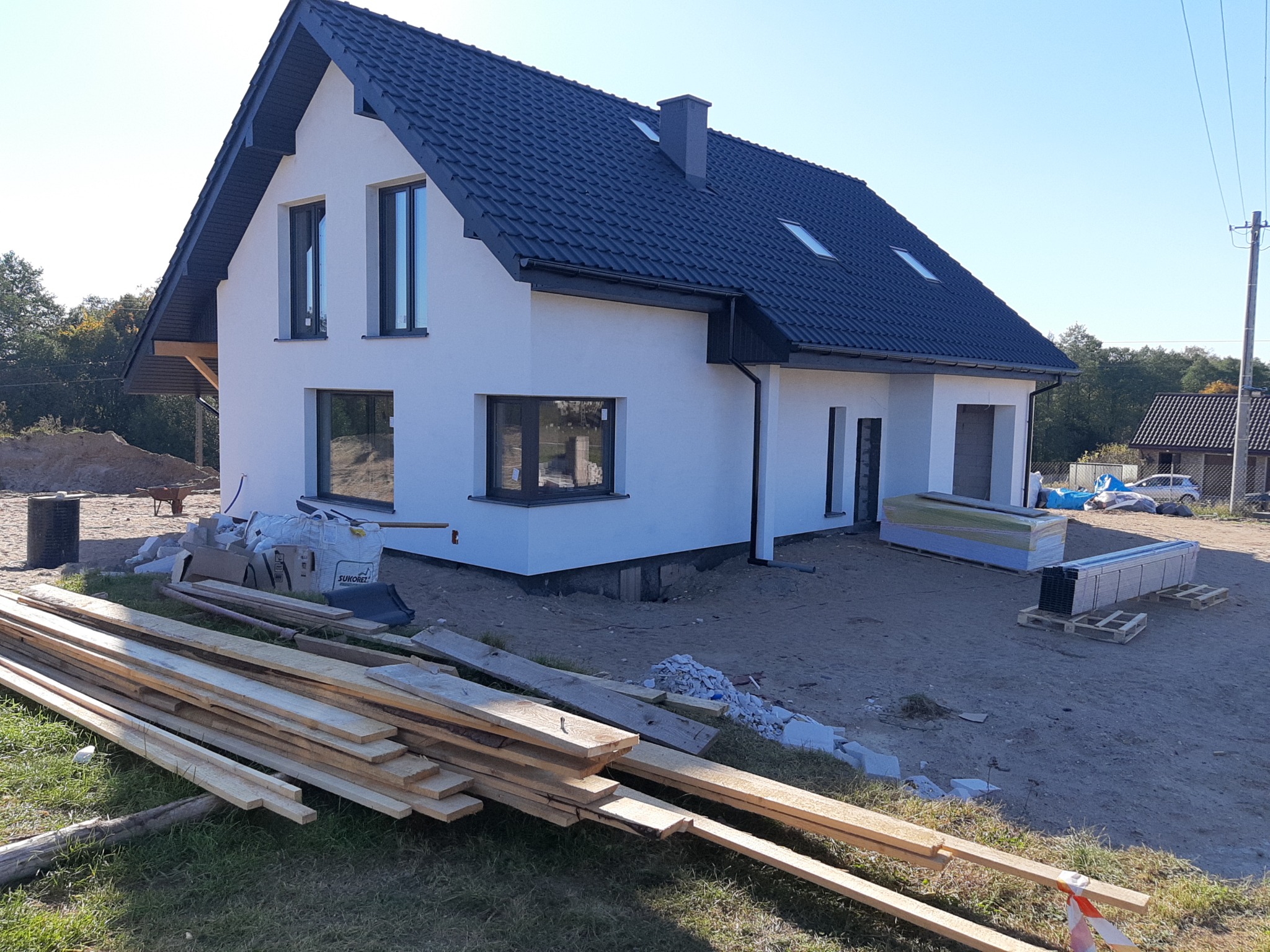 Nowoczesny dom jednorodzinny z białą elewacją i ciemną dachówką, widok z zewnątrz, z materiałami budowlanymi leżącymi na ziemi, sugerującymi trwające prace wykończeniowe.