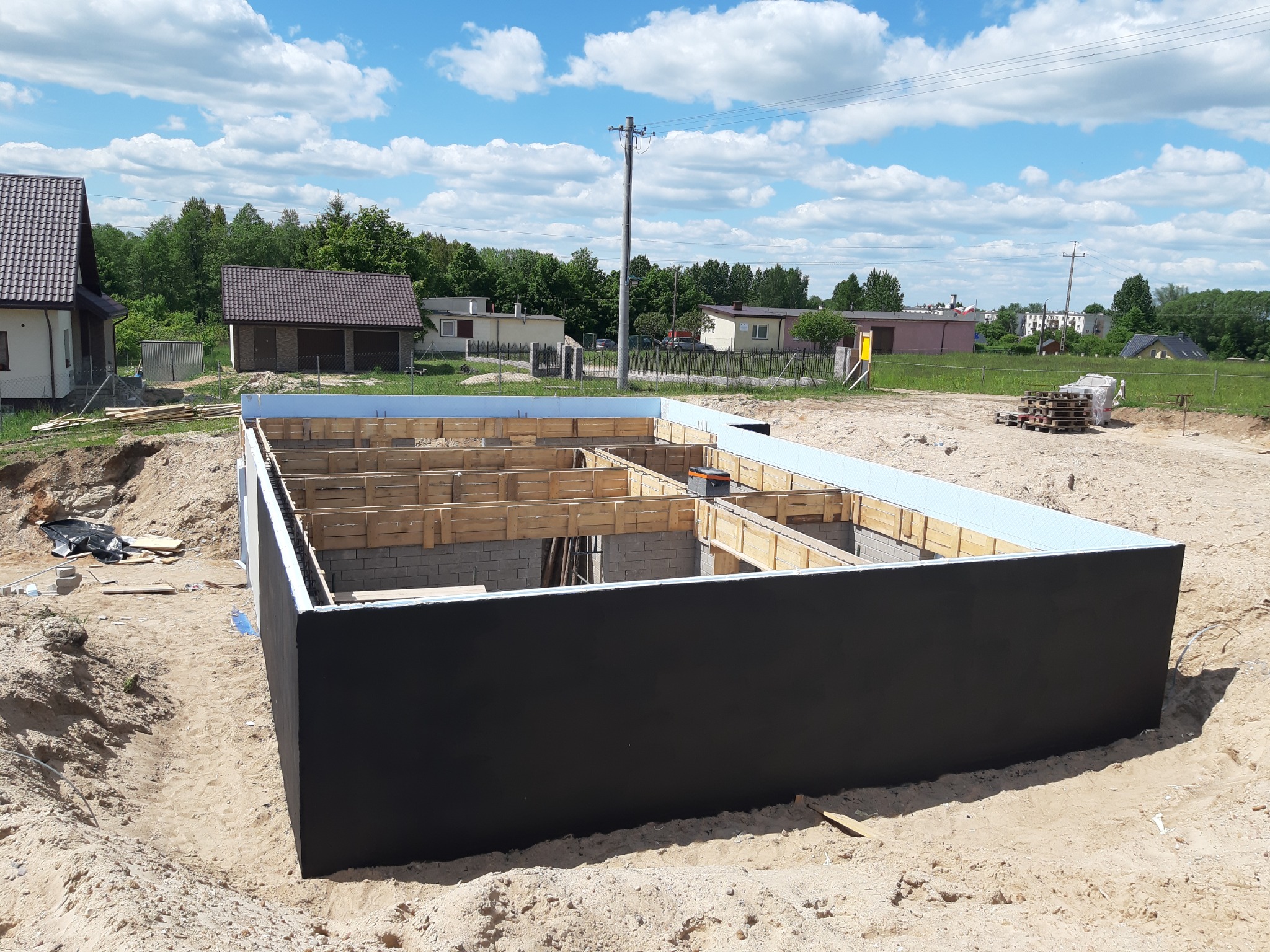 Fundament domu w trakcie budowy, z widocznym szalunkiem i izolacją, na tle piaszczystego terenu i zabudowań jednorodzinnych pod błękitnym niebem.