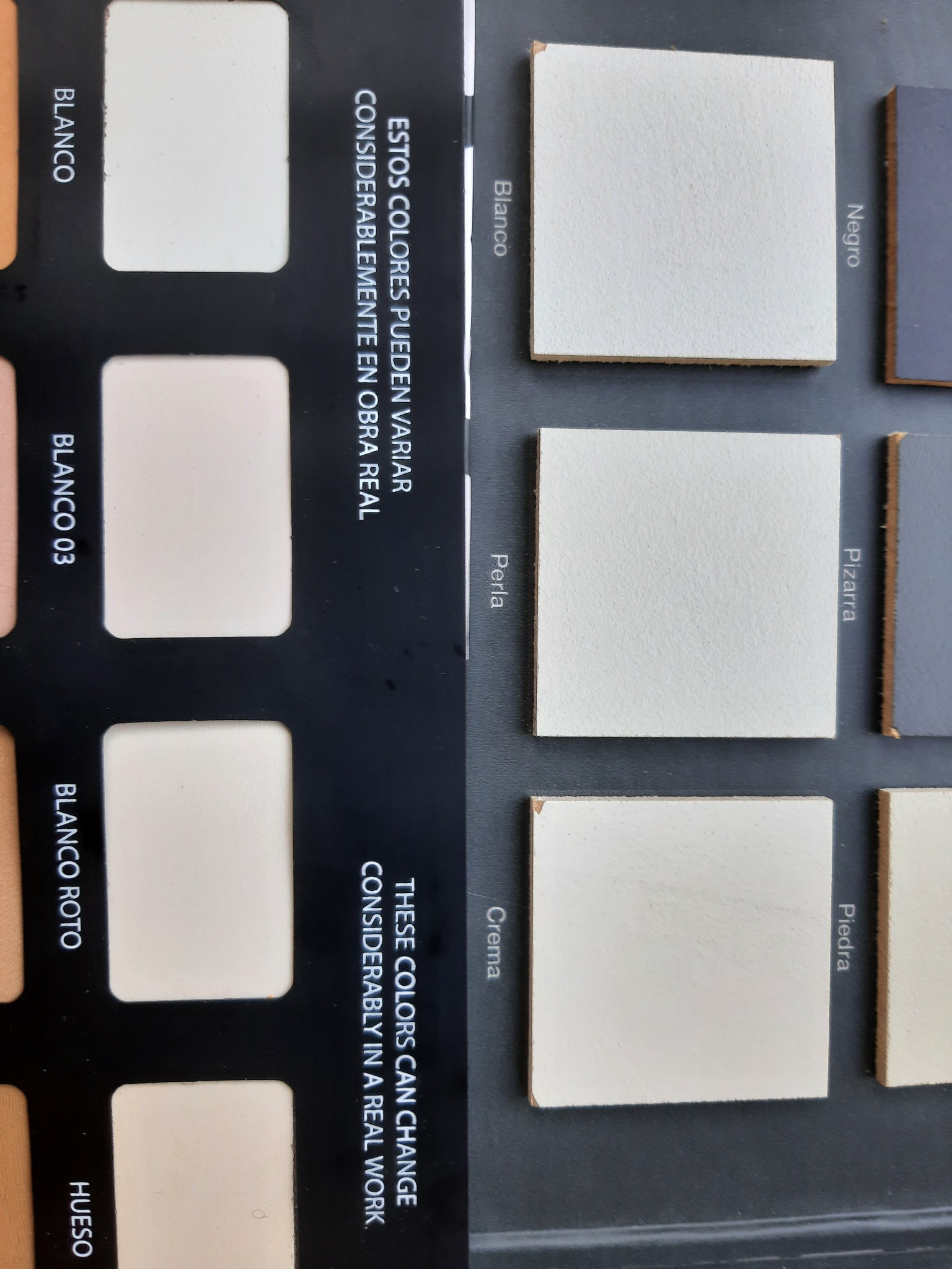 Paleta próbek kolorystycznych z kwadratowymi płytkami w odcieniach bieli, szarości i beżu, z nazwami kolorów w języku hiszpańskim i angielskim informującymi o możliwych różnicach w odcieniach...