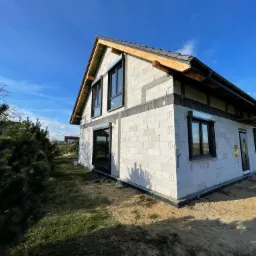 Surowy stan budynku jednorodzinnego z zamontowanymi czarnymi oknami, widoczne bloczki z betonu komórkowego, drewniana konstrukcja dachu i częściowo ułożona dachówka, w tle błękitne niebo.