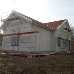 Boike Building Krzysztof Boike - Otynkowany dom jednorodzinny z czerwoną dachówką, otoczony rusztowaniami. Okna zabezpieczone folią. Widoczne świeże nasypy ziemi.