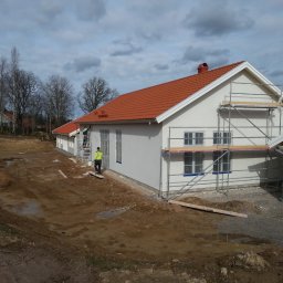 Boike Building Krzysztof Boike - Biały dom z czerwoną dachówką w trakcie prac wykończeniowych. Widoczne rusztowania, pracownik w żółtej kamizelce i teren wokół domu w stanie surowym.