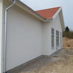 Boike Building Krzysztof Boike - Boczny widok otynkowanego domu z czerwoną dachówką, widoczne okna, rynna i fragment terenu z pracownikiem w oddali. Jasna elewacja, spokojna kolorystyka.