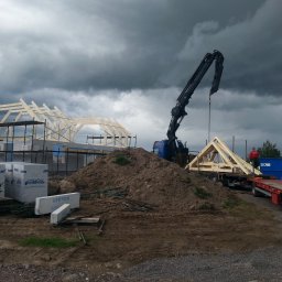 Boike Building Krzysztof Boike - Montaż drewnianej konstrukcji domu szkieletowego za pomocą dźwigu na tle pochmurnego nieba. Widoczne elementy konstrukcyjne i materiały budowlane.