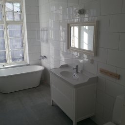 Boike Building Krzysztof Boike - Jasna łazienka z wanną wolnostojącą, białą umywalką z szafką i szarą podłogą. Ściany wyłożone białymi, prostokątnymi płytkami. Widoczne okno z naturalnym światłem.