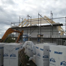 Boike Building Krzysztof Boike - Budowa domu z bloczków Solbet, widoczne rusztowania, dźwig i drewniana konstrukcja dachu na tle pochmurnego nieba. Materiały budowlane na pierwszym planie.