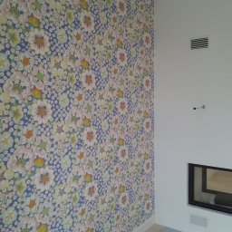 Boike Building Krzysztof Boike - Ściana z tapetą w kwiaty i biały kominek z szybą. Widoczna kratka wentylacyjna i fragment podłogi. Wykończenie wnętrza w jasnych barwach.