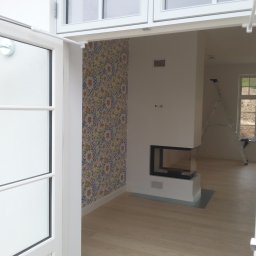 Boike Building Krzysztof Boike - Otwarty widok na jasne wnętrze z kominkiem i wzorzystą tapetą, widziane przez białe, uchylone drzwi balkonowe. W tle drabina oparta o ścianę.