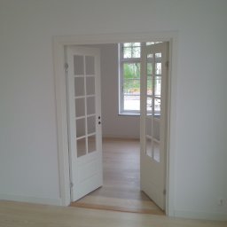 Boike Building Krzysztof Boike - Białe, przeszklone drzwi dwuskrzydłowe, otwarte do wnętrza jasnego pokoju z drewnianą podłogą. Widok na okno z zielenią za nim. Minimalistyczne wykończenie.