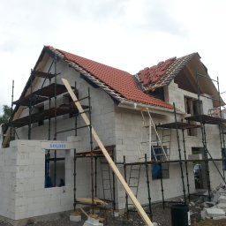Boike Building Krzysztof Boike - Budowa domu z białych bloczków, częściowo zadaszonego czerwoną dachówką. Widoczne rusztowania, drabina i resztki materiałów budowlanych. Etap surowy otwarty.