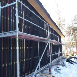 Boike Building Krzysztof Boike - Ciemna elewacja domu szkieletowego w trakcie montażu, widoczna spod rusztowania. Elementy konstrukcyjne i ocieplenie częściowo odsłonięte. Śnieg na ziemi.