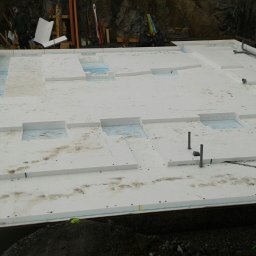 Boike Building Krzysztof Boike - Fundament z izolacją termiczną w postaci białych płyt styropianowych, widoczne wystające rury instalacyjne. Prace budowlane w toku, teren nierówny.