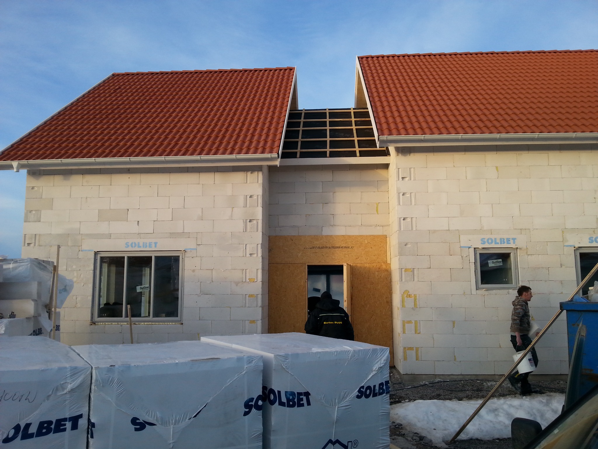 Budowa domu z białych bloczków Solbet z czerwoną dachówką. Widoczne okna, wejście zabezpieczone płytą OSB i pracownik z wiadrem. Częściowo ułożone pokrycie dachu.