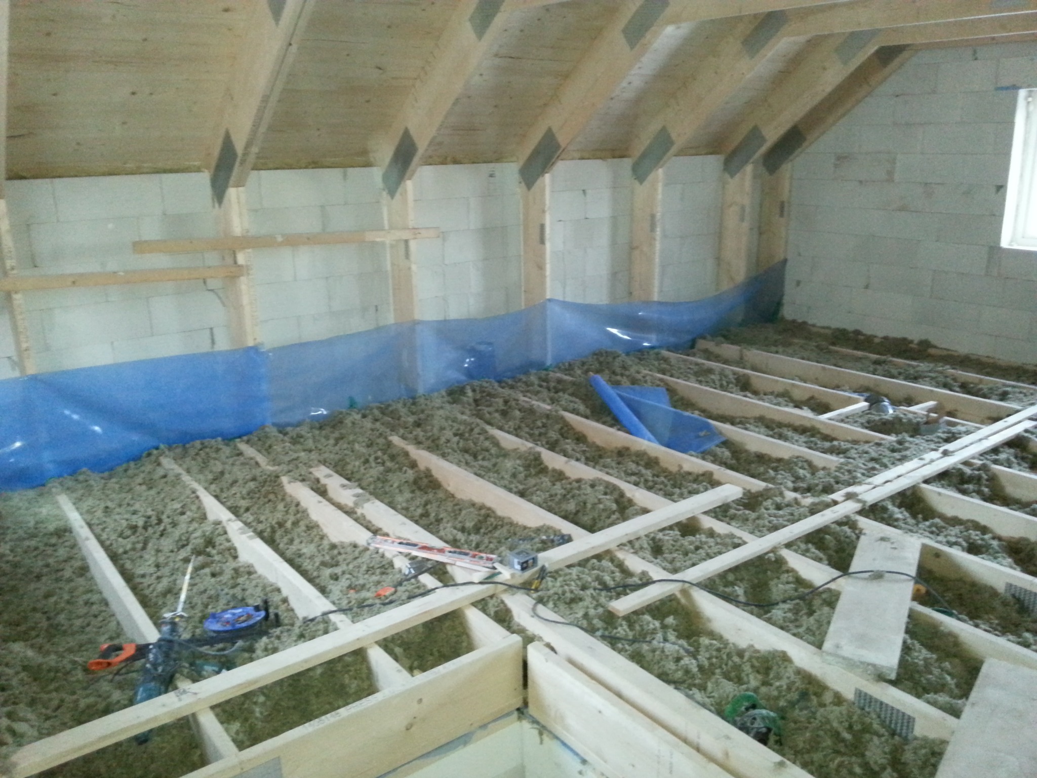 Ocieplenie poddasza wełną mineralną, widoczne drewniane legary i narzędzia pracy. Niebieska folia zabezpieczająca przy ścianie z bloczków.