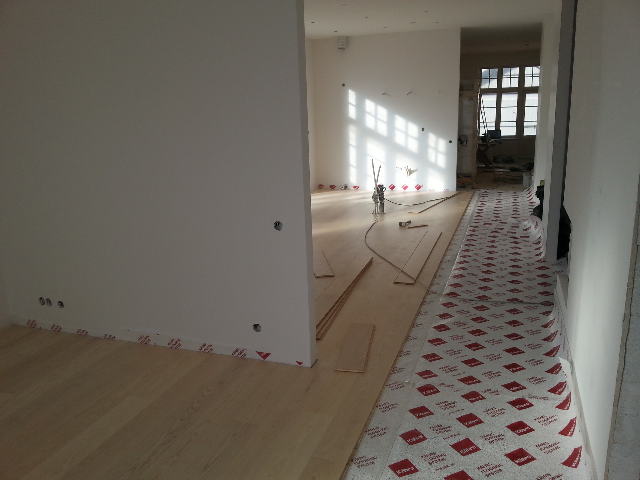 Układanie jasnej drewnianej podłogi w przestronnym wnętrzu. Część podłogi zabezpieczona folią ochronną z czerwonym nadrukiem. Widoczne narzędzia i przygotowane deski.