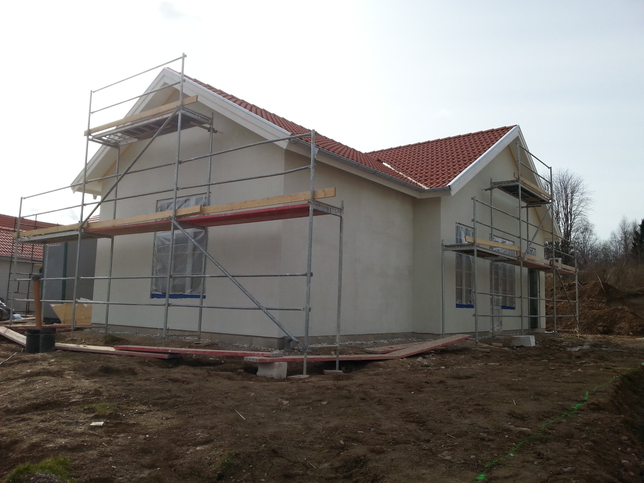 Otynkowany dom jednorodzinny z czerwoną dachówką, otoczony rusztowaniami. Okna zabezpieczone folią. Widoczne świeże nasypy ziemi.